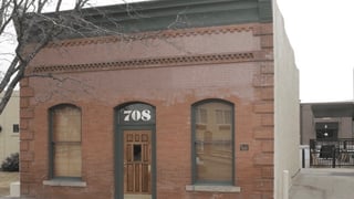 Exterior view of a brick commercial building with a wooden door featuring the number 708 above it, and two arched windows with blinds.