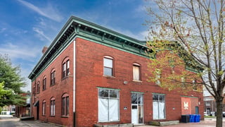 Exterior view of a red brick commercial building with green trim and multiple windows, under a blue sky with some trees in the foreground and background.