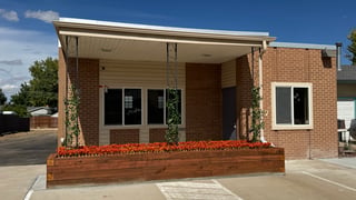 The exterior of a single-story brick commercial building with a covered entrance supported by decorative metal posts, a wooden planter box with orange flowers, and a concrete driveway.