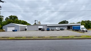 Exterior view of a gray industrial building complex with a gravel parking lot, several cars, a large blue semi-truck at a loading dock, and surrounding trees under an overcast sky.